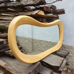 May include: A light-colored, wooden mirror with a unique, wavy frame. The mirror has a rectangular shape with rounded corners and is made of light-colored wood. The frame is thick and has a smooth, curved design. The mirror is displayed on a pile of wooden planks.