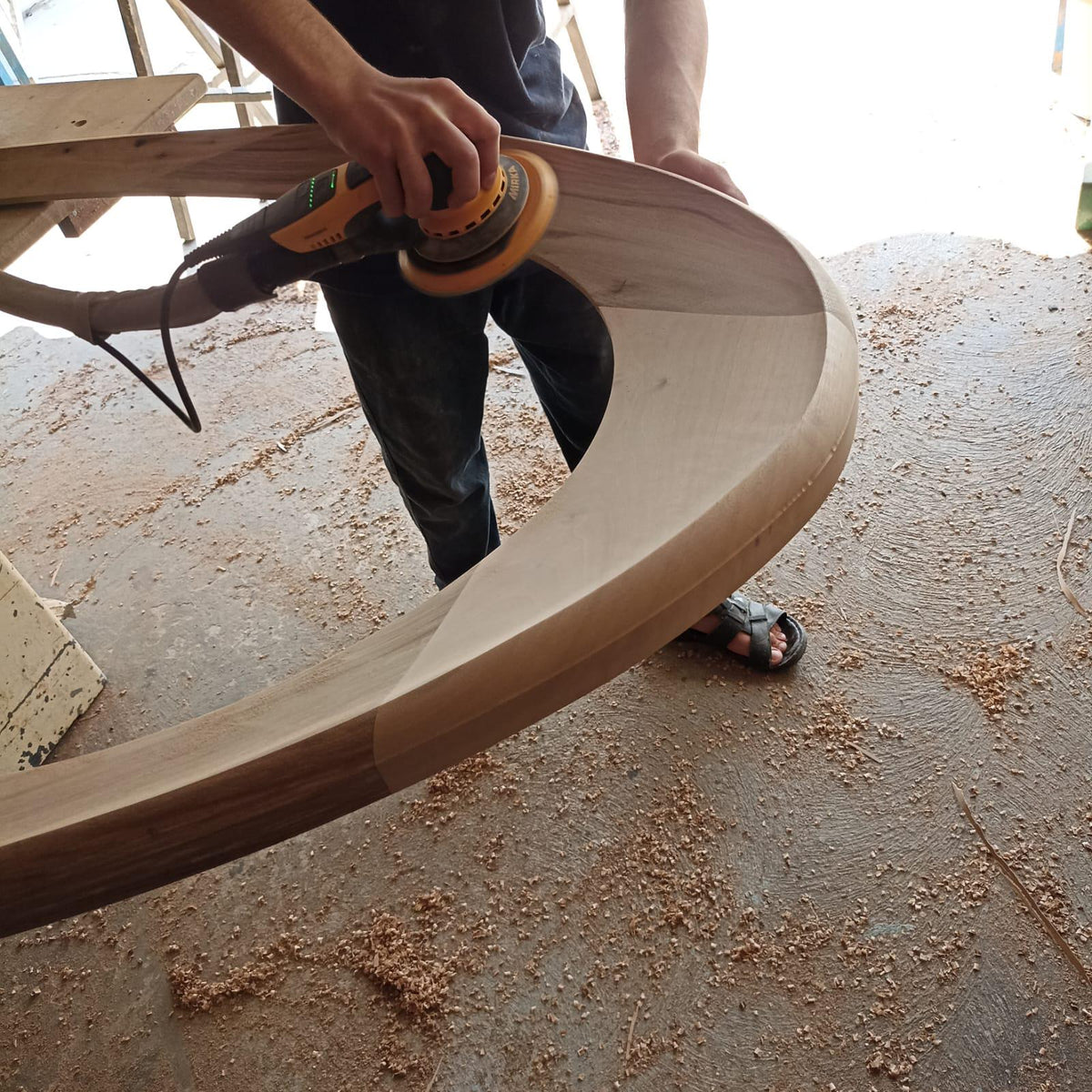 May include: A craftsman sands a curved wooden furniture piece with a power sander. The wood is light brown, and the sander is yellow and black. Sawdust covers the concrete floor.