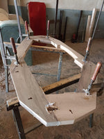 May include: A partially constructed wooden frame, likely for furniture, held together with clamps. The light-colored wood is being assembled on a work table, with a red chair visible in the background. The workshop setting suggests a focus on woodworking and craftsmanship.