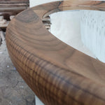 May include: Close-up of a curved wooden frame, likely part of a mirror or decorative piece. The wood has a rich, dark brown color with visible grain patterns. The frame curves smoothly, suggesting a modern design.