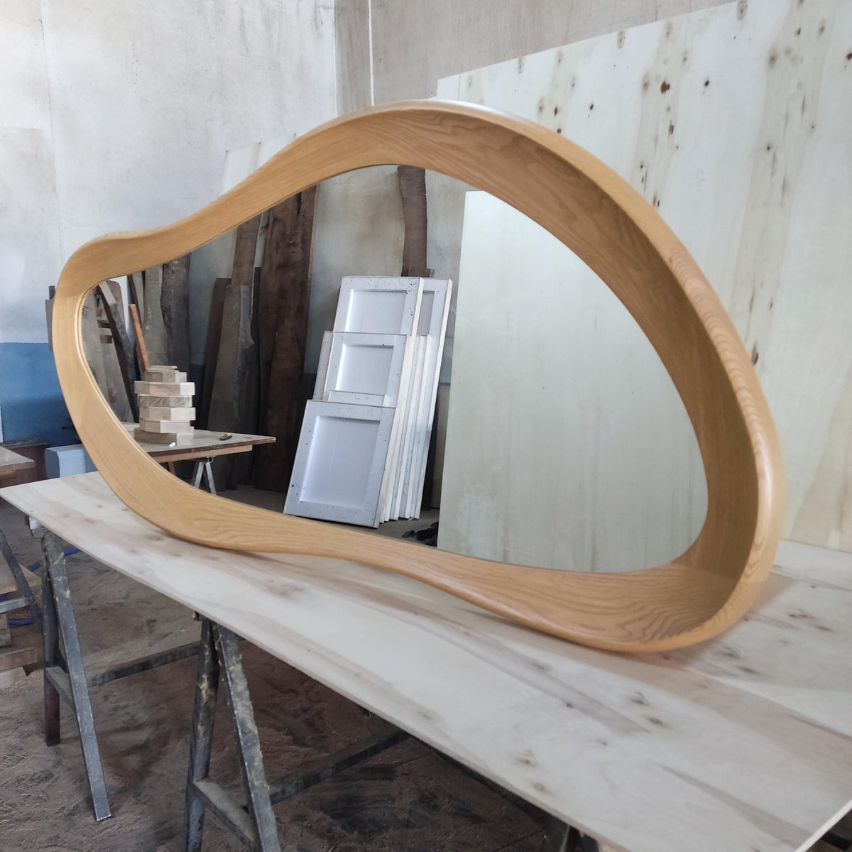 May include: A large, organically shaped mirror with a light brown wooden frame. The frame has a smooth, curved design. The mirror is resting on a light-colored wooden surface, reflecting the surrounding workshop environment.
