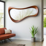 May include: A large, organically shaped wall mirror with a warm brown wooden frame. The mirror reflects a view of tree branches against a bright sky. The mirror is mounted on a white wall above a red sofa.
