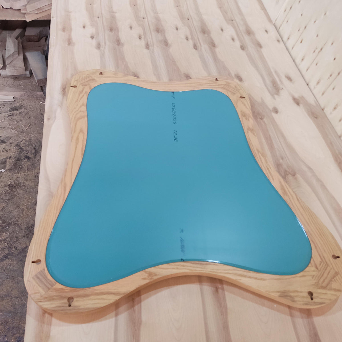 May include: A unique, irregularly shaped mirror with a teal-colored reflective surface. The mirror is framed by a light-colored, natural wood border. Mounting hardware is visible on the back of the frame. The mirror is likely for home decor.