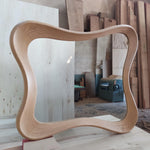 May include: A light-colored wooden mirror with a unique, wavy frame. The frame is made of light-colored wood and has a smooth, curved design. The mirror is rectangular with rounded corners. The mirror is displayed in a workshop setting.