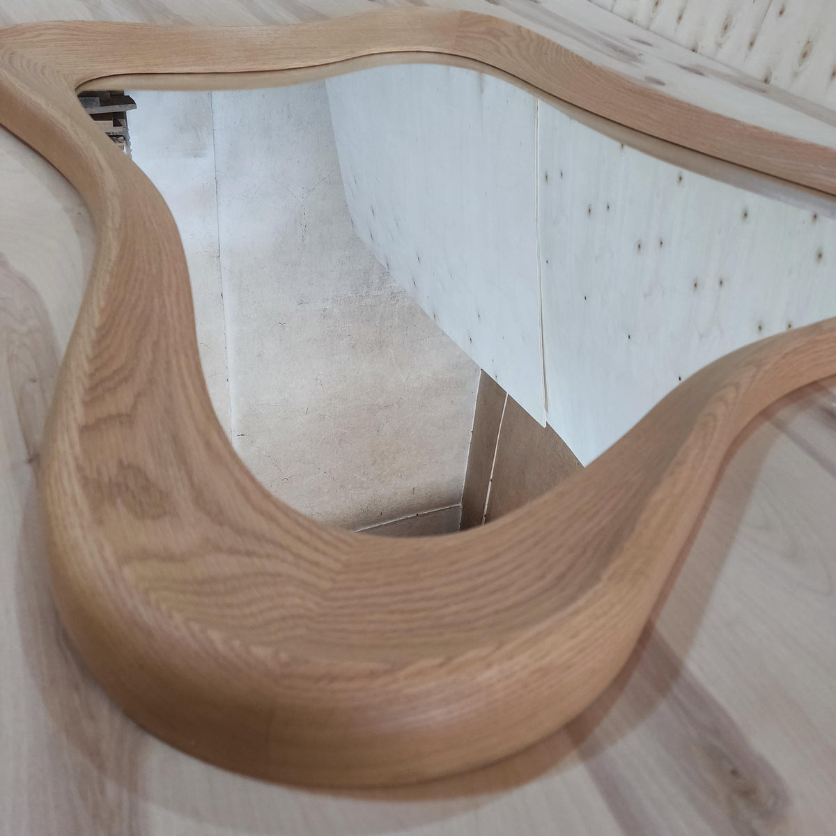 May include: A large, wavy-edged mirror with a light brown wooden frame. The frame has a smooth, curved design. The mirror reflects the surrounding environment, including a light-colored wall and wooden floor.
