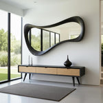 May include: A modern entryway featuring a large, black, organically shaped mirror. Below the mirror is a black and wood-toned console table with three drawers. Two white vases and a black vase sit on the console. A gray rug is on the floor.