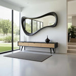 May include: A modern entryway featuring a large, black, organically shaped mirror above a black and wood-toned console table. The console has three drawers and sits on angled legs. A gray rug and decorative vases complete the space.