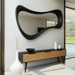 May include: A large, black, organically shaped mirror hangs above a modern wooden console table. The console has two drawers with light wood fronts and a dark top. Decorative white vases and a dark bowl sit on the console.