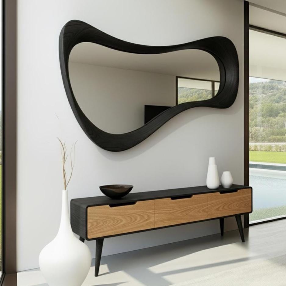 May include: A large, black, organically shaped mirror hangs above a modern wooden console table. The console has two drawers with light wood fronts and a dark top. Decorative white vases and a dark bowl sit on the console.
