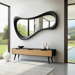 May include: A modern living room features a large, black, wavy-edged mirror mounted on a white wall. Below, a wooden cabinet with three drawers and black legs holds white vases and a dark bowl. A white rug and large windows complete the scene.