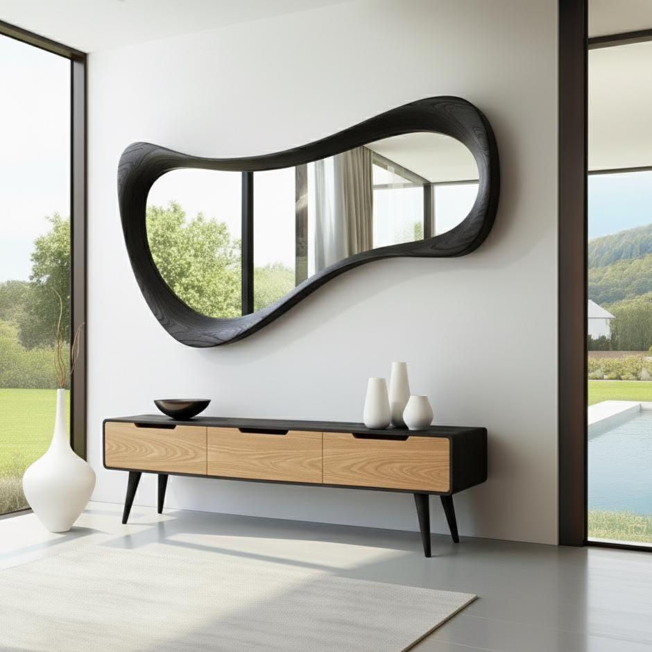 May include: A modern living room features a large, black, wavy-edged mirror mounted on a white wall. Below, a wooden cabinet with three drawers and black legs holds white vases and a dark bowl. A white rug and large windows complete the scene.