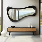 May include: A modern, black-framed mirror with an organic shape hangs above a wooden console table. The console has three drawers with a light wood grain finish and black accents. White vases and a black bowl are on the console.