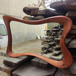 May include: A large, wavy-edged mirror with a reddish-brown wooden frame. The frame has a smooth, polished finish and a unique, flowing design. The mirror is displayed outdoors, resting on a stack of wooden planks.