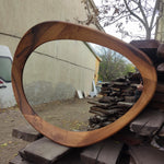 May include: A large, oval-shaped wooden mirror with a natural wood grain finish. The frame is thick and curved, creating a unique, organic shape. The mirror reflects the surrounding environment, showcasing its reflective surface.