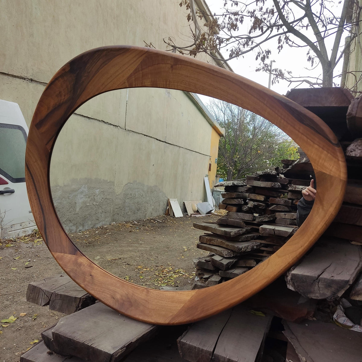 May include: A large, oval-shaped wooden mirror with a natural wood grain finish. The frame is thick and curved, creating a unique, organic shape. The mirror reflects the surrounding environment, showcasing its reflective surface.
