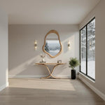 May include: A modern entryway featuring a wooden console table with a curved base, topped with a vase, books, and a small clock. Above the table hangs an organic-shaped wooden mirror. The room has light-colored walls and flooring, with a large window overlooking a snowy landscape.