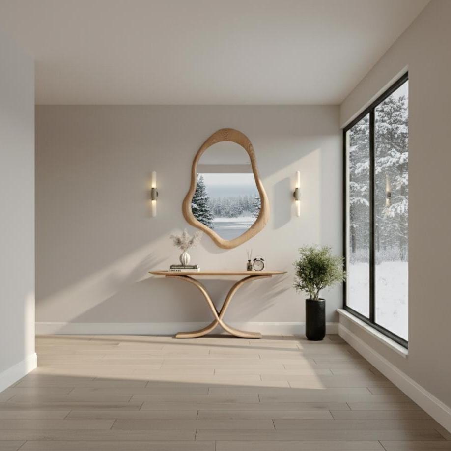 May include: A modern entryway featuring a wooden console table with a curved base, topped with a vase, books, and a small clock. Above the table hangs an organic-shaped wooden mirror. The room has light-colored walls and flooring, with a large window overlooking a snowy landscape.