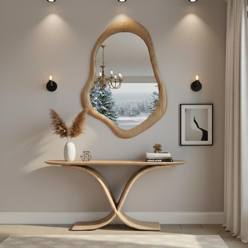 May include: A wooden console table with a unique curved base, topped with a decorative vase, geometric object, and books. Above, a large, organically shaped wooden mirror reflects a snowy landscape. Two black sconces flank a framed abstract print.