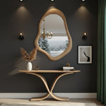 May include: A wooden console table with a unique, curved base supports a vase of dried flowers, a small geometric object, and books. Above, a large, organically shaped wooden mirror reflects a snowy landscape. The wall is dark gray with sconces and a framed print.