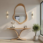 May include: A wooden console table with a unique curved base, topped with a vase, books, and a small clock. A large, organically shaped wooden mirror hangs above. Two wall sconces flank the mirror. A potted plant sits to the right.