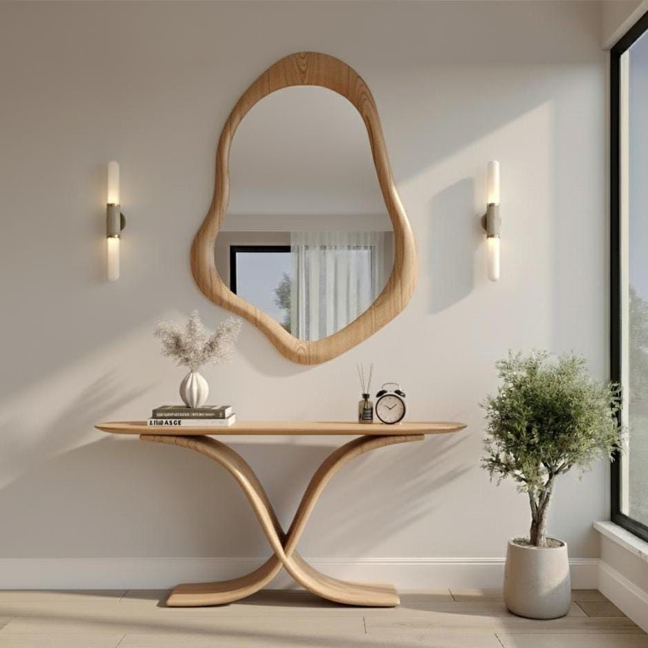 May include: A wooden console table with a unique curved base, topped with a vase, books, and a small clock. A large, organically shaped wooden mirror hangs above. Two wall sconces flank the mirror. A potted plant sits to the right.