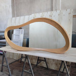 May include: A large, organically shaped wooden frame, possibly for a mirror or artwork, is displayed on a light-colored wooden surface. The frame has a smooth, curved design and a natural wood grain finish. The piece is set against a backdrop of wooden panels.