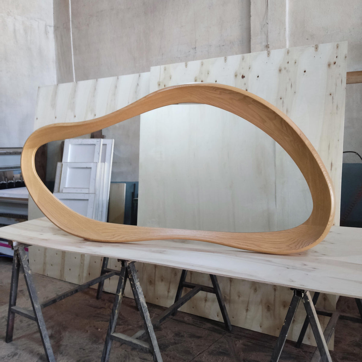 May include: A large, organically shaped wooden frame, possibly for a mirror or artwork, is displayed on a light-colored wooden surface. The frame has a smooth, curved design and a natural wood grain finish. The piece is set against a backdrop of wooden panels.