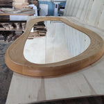 May include: A large, organically shaped mirror with a light brown wooden frame. The frame has a smooth, rounded design. The mirror reflects the surrounding workshop environment, including stacks of wood and other materials.