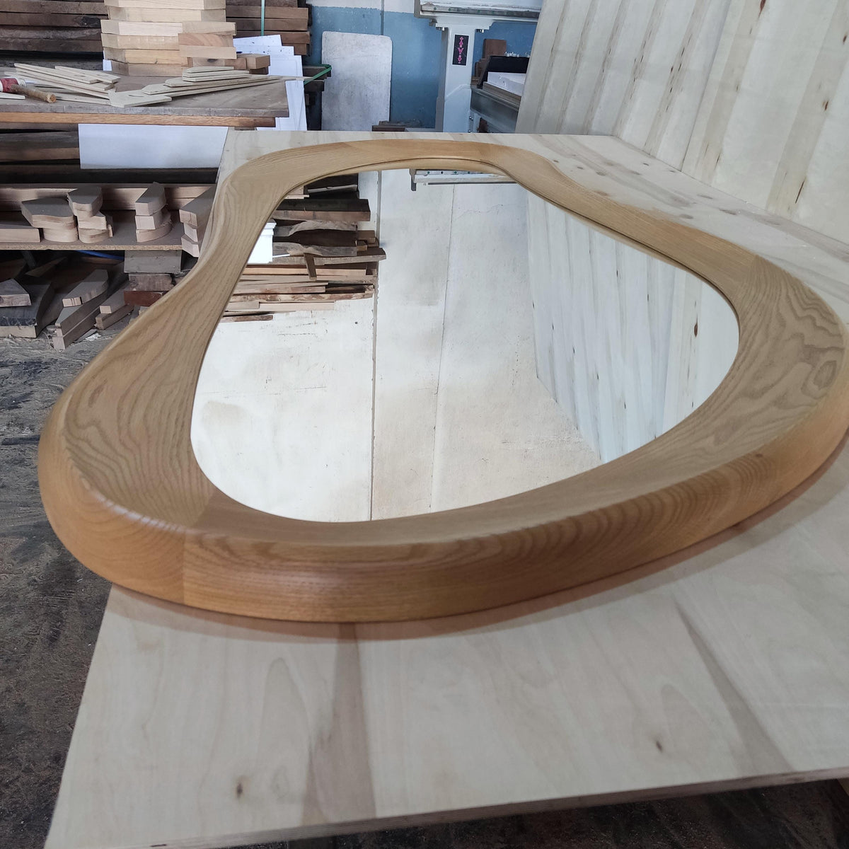 May include: A large, organically shaped mirror with a light brown wooden frame. The frame has a smooth, rounded design. The mirror reflects the surrounding workshop environment, including stacks of wood and other materials.