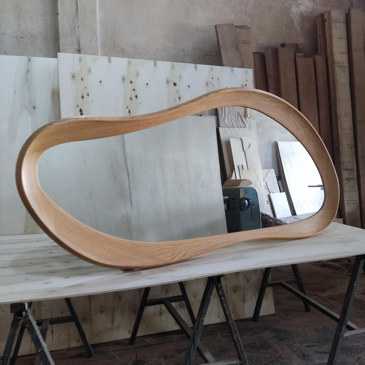 May include: A large, organically shaped mirror with a light brown wooden frame. The mirror is displayed on a light-colored wooden surface, showcasing its unique, flowing design. The frame's smooth curves and natural wood grain add a touch of modern elegance.