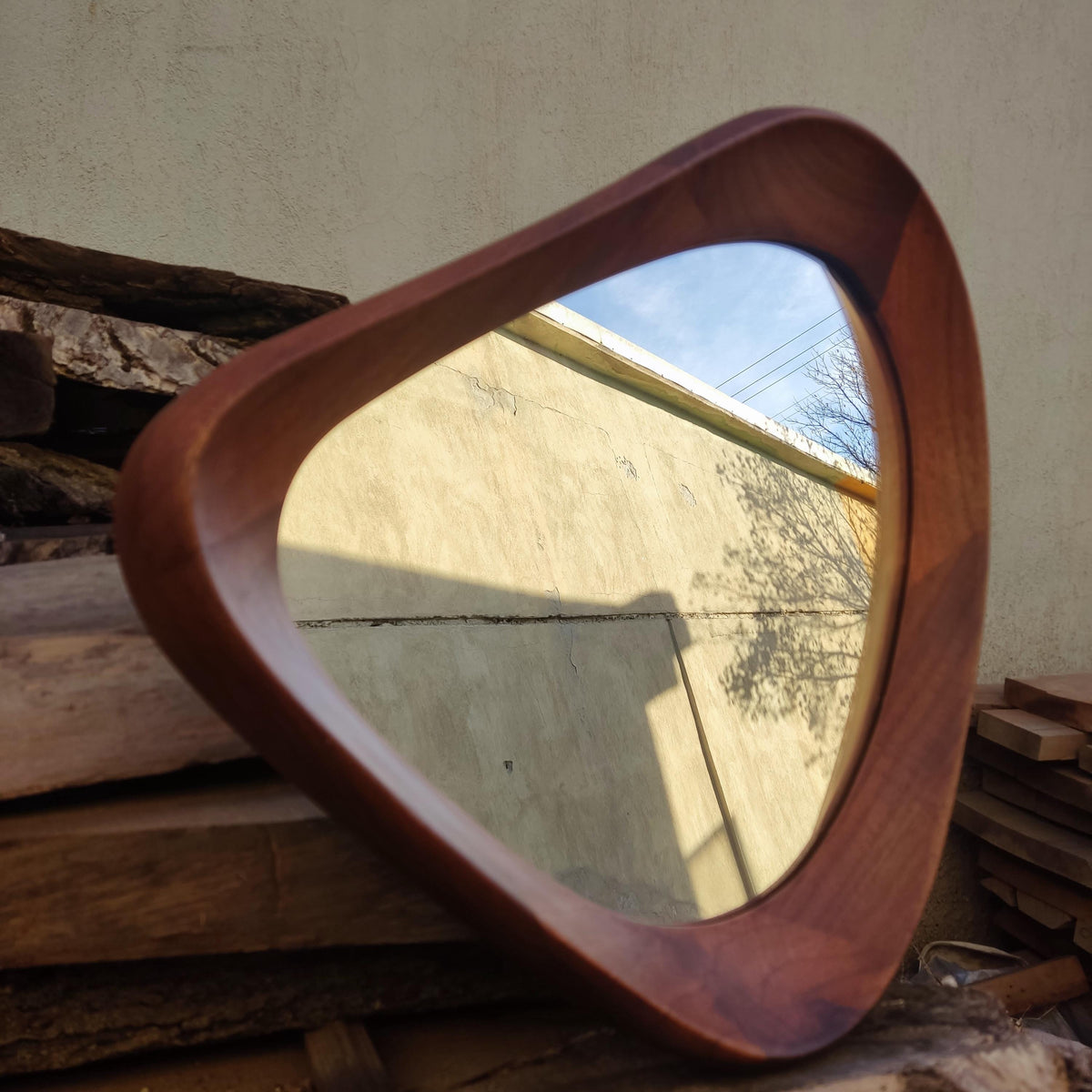May include: A mid-century modern style mirror with a unique, rounded triangular shape. The frame is made of dark brown wood, and the mirror reflects a view of a blue sky and a tree. The mirror is set against a neutral-colored wall.