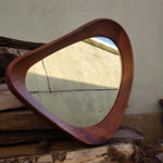 May include: A mid-century modern style mirror with a unique, rounded triangular shape. The frame is made of dark brown wood, and the mirror reflects the surrounding environment. The mirror is a decorative item.