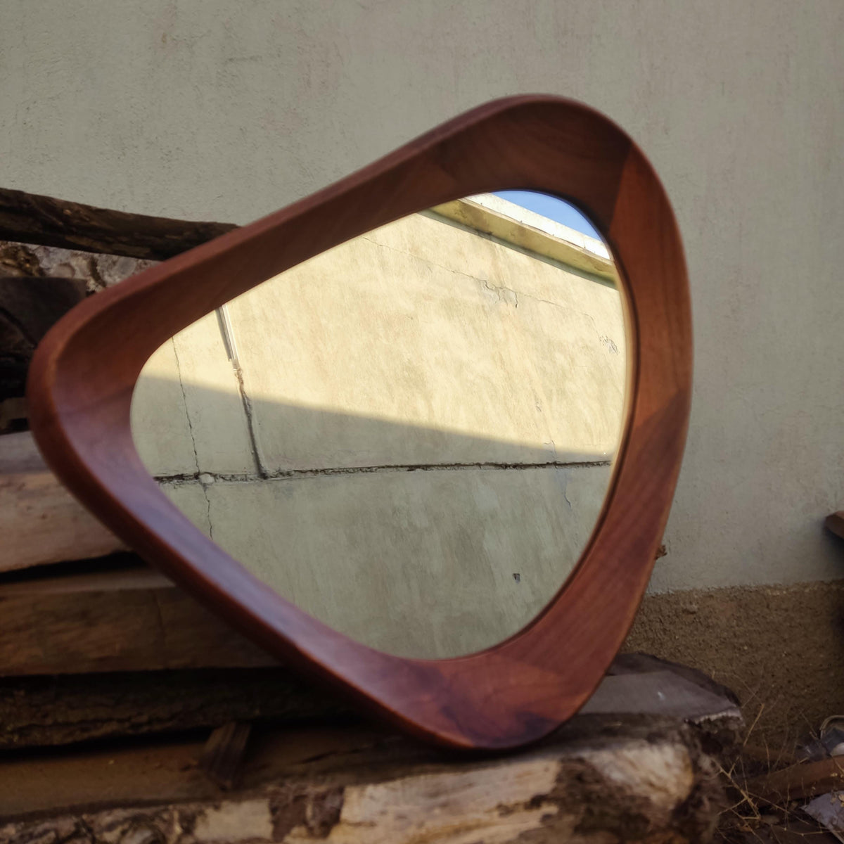 May include: A mid-century modern style mirror with a unique, rounded triangular shape. The frame is made of dark brown wood, and the mirror reflects the surrounding environment. The mirror is a decorative item.