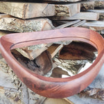 May include: A handcrafted, teardrop-shaped wooden mirror with a rich, reddish-brown finish. The mirror's frame is smoothly curved, and the reflective surface is visible within. The mirror is set against a backdrop of stacked wooden planks.