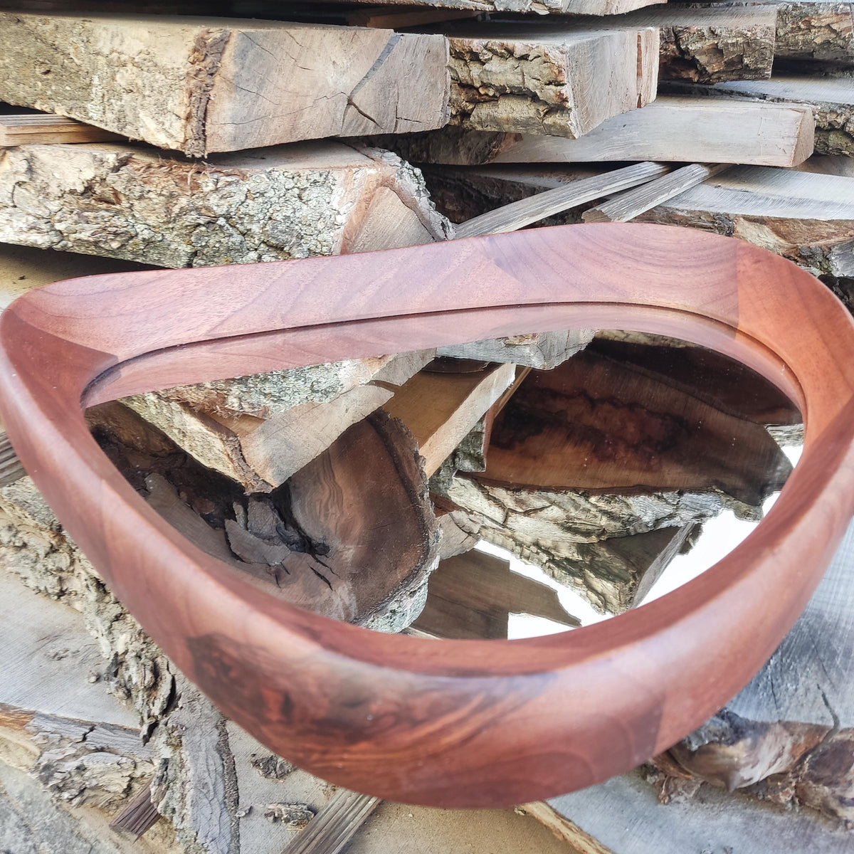 May include: A handcrafted, teardrop-shaped wooden mirror with a rich, reddish-brown finish. The mirror's frame is smoothly curved, and the reflective surface is visible within. The mirror is set against a backdrop of stacked wooden planks.