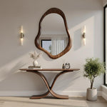 May include: A modern entryway featuring a large, organically shaped wooden mirror above a matching console table. The table has a unique curved base. Decorative items include a vase with dried flowers, a book, a small clock, and a potted plant.