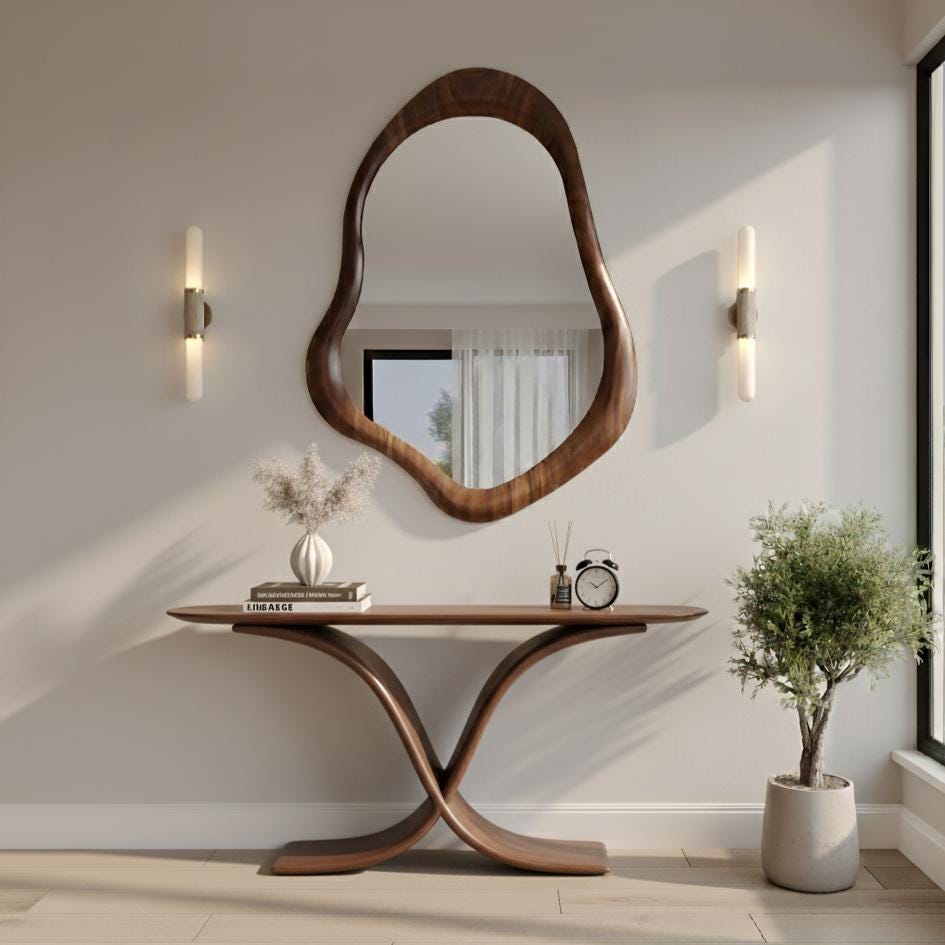 May include: A modern entryway featuring a large, organically shaped wooden mirror above a matching console table. The table has a unique curved base. Decorative items include a vase with dried flowers, a book, a small clock, and a potted plant.