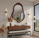 May include: A modern entryway featuring a large, organically shaped wooden mirror. Below, a floating wooden console with a bench and shoe rack. Decorative elements include pampas grass, a floor lamp, and wall sconces.