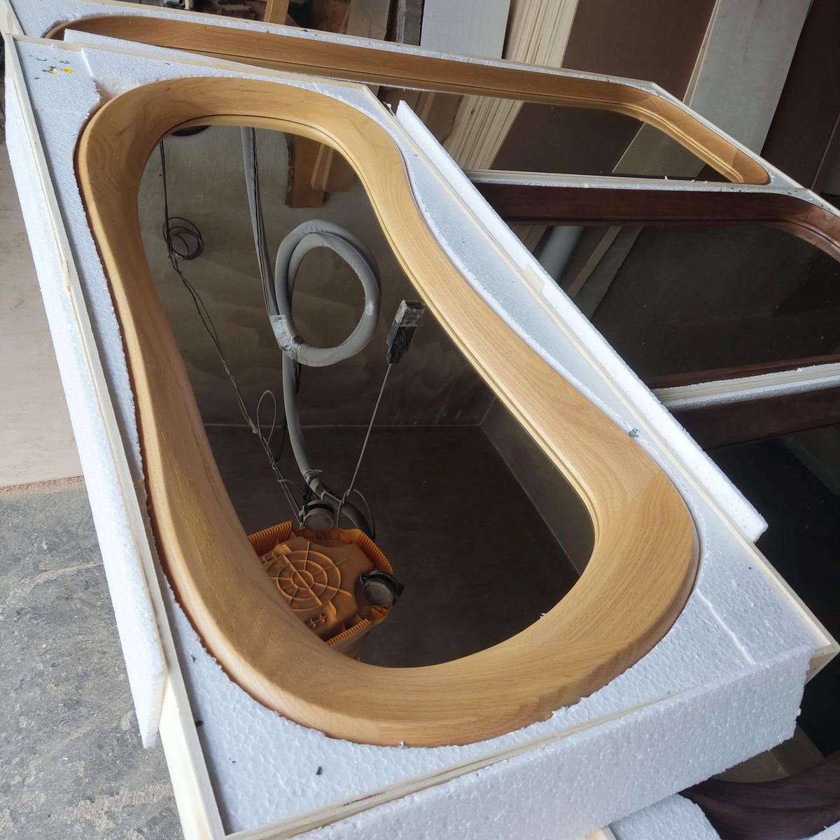 May include: A large, light-colored wooden framed mirror with a unique, curved design. The mirror is encased in white protective foam packaging. Electrical wires and a yellow component are visible inside the frame.