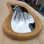 May include: A uniquely shaped mirror with a light wood frame. The mirror's reflective surface shows a cloudy sky and tree branches. The frame has a smooth, curved design, adding a modern touch to the decor.