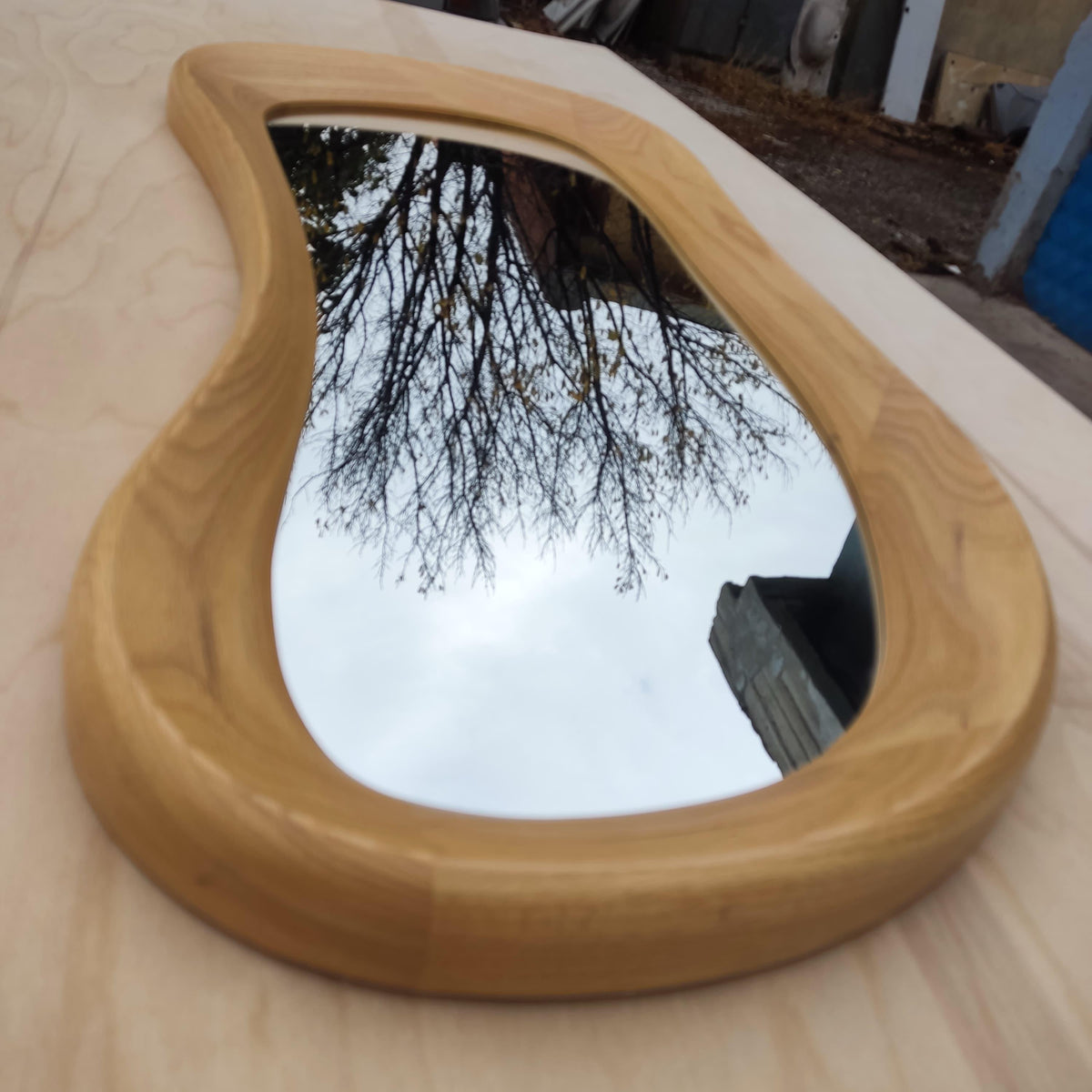 May include: A uniquely shaped mirror with a light wood frame. The mirror's reflective surface shows a cloudy sky and tree branches. The frame has a smooth, curved design, adding a modern touch to the decor.