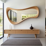 May include: A modern, organic-shaped wooden mirror hangs above a matching wooden console table. The table has three drawers and tapered legs. Two white vases and a dark brown vase sit on the table. A gray rug is on the floor.