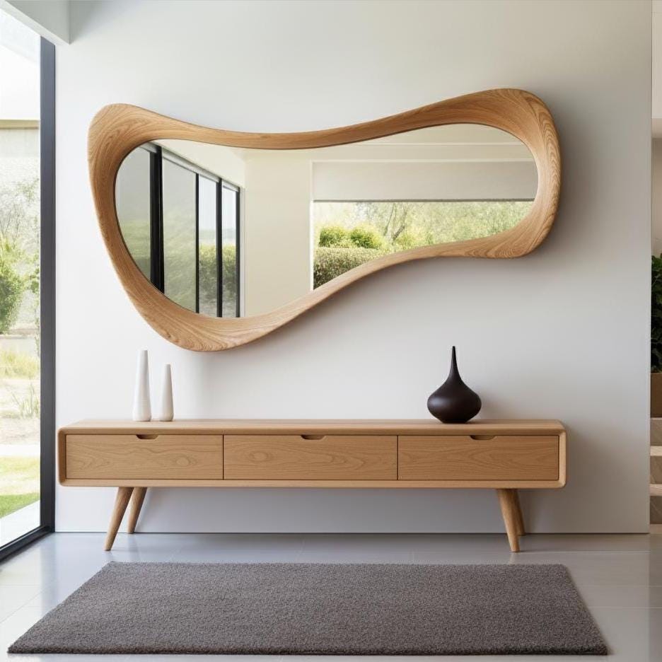 May include: A modern, organic-shaped wooden mirror hangs above a matching wooden console table. The table has three drawers and tapered legs. Two white vases and a dark brown vase sit on the table. A gray rug is on the floor.