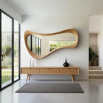 May include: A modern, organic-shaped wooden mirror hangs above a light wood console with three drawers. Two white vases and a dark brown vase sit on the console. A gray rug lies on the floor. The room has large windows and a minimalist design.