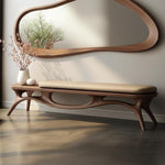 May include: A modern wooden bench with a tan cushion, featuring a unique, curved design. Above, a large, organically shaped wooden mirror hangs on the wall. The scene is complemented by a vase of flowers and a small decorative object on the bench.