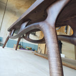 May include: A low-angle view of a handcrafted wooden table. The table features a dark brown finish with organically shaped legs and supports. The design showcases a modern aesthetic with smooth, curved lines and a sturdy construction.