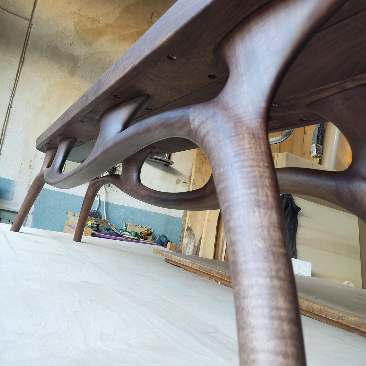 May include: A low-angle view of a handcrafted wooden table. The table features a dark brown finish with organically shaped legs and supports. The design showcases a modern aesthetic with smooth, curved lines and a sturdy construction.