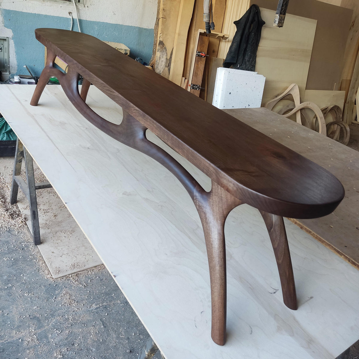 May include: A long, dark brown wooden bench with a curved design. The bench has four legs and a smooth, polished surface. The legs are curved and have a unique shape. The bench is approximately 72 inches long.