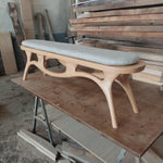 May include: A light-colored wooden bench with a curved design and a cushioned seat. The bench has four legs and a long, rectangular seat cushion. The wood has a natural finish, and the cushion is a light gray color. The bench is likely for indoor use.
