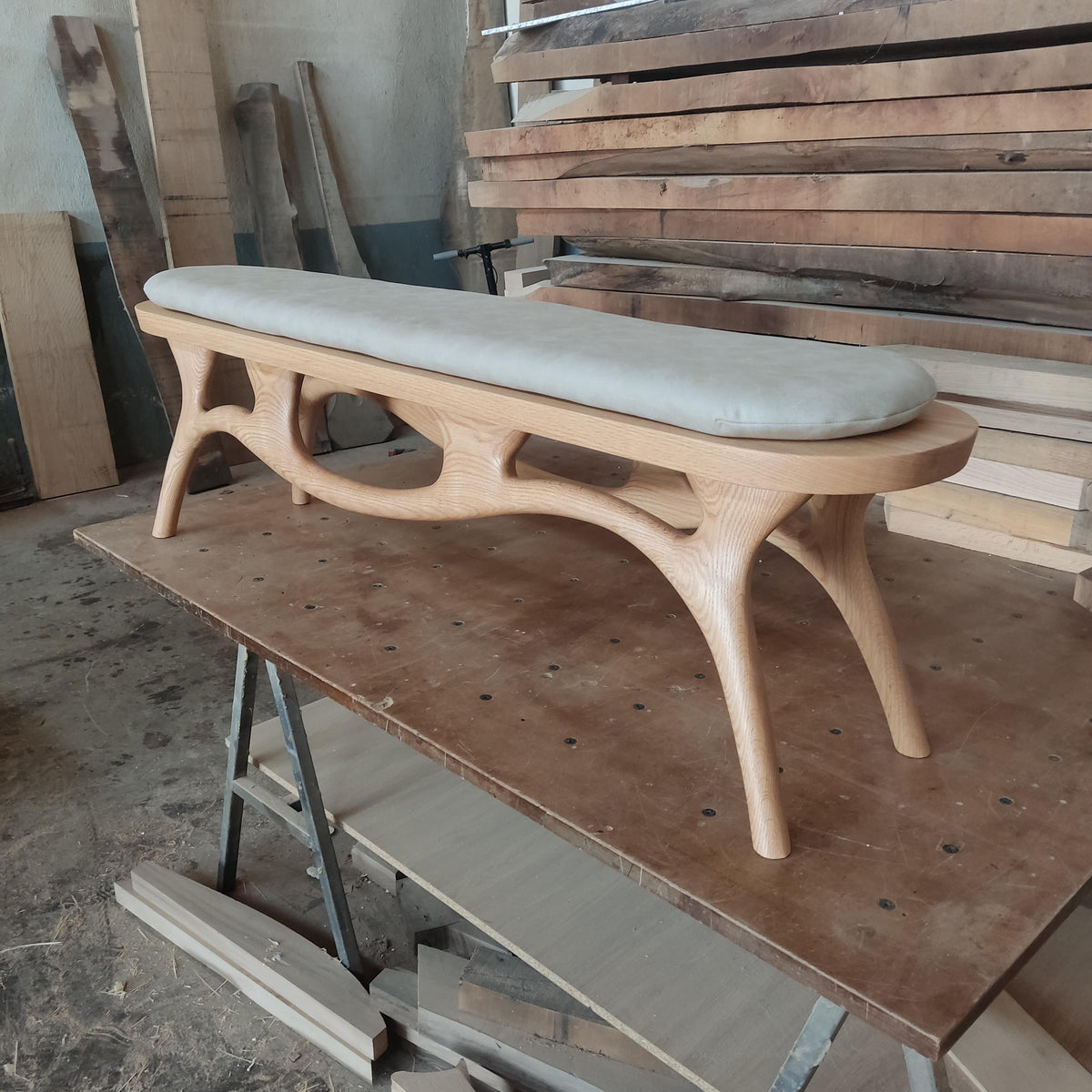 May include: A light-colored wooden bench with a curved design and a cushioned seat. The bench has four legs and a long, rectangular seat cushion. The wood has a natural finish, and the cushion is a light gray color. The bench is likely for indoor use.
